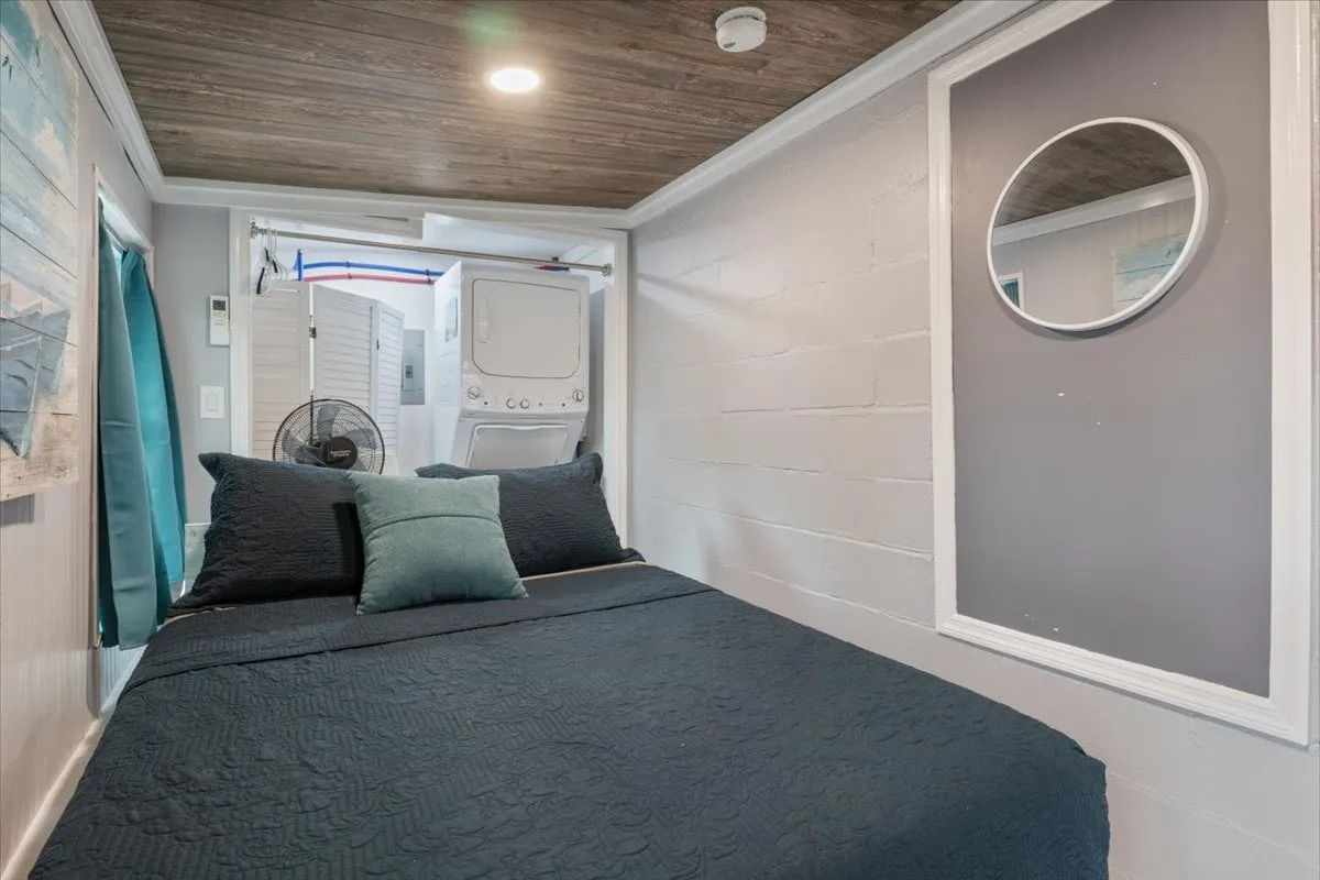 An intimate bedroom scene with a dark-colored bed, decorative pillow, a round mirror above, and a view of a laundry area in the background. The mood is comfortable and homely, evoking a sense of retreat and relaxation.