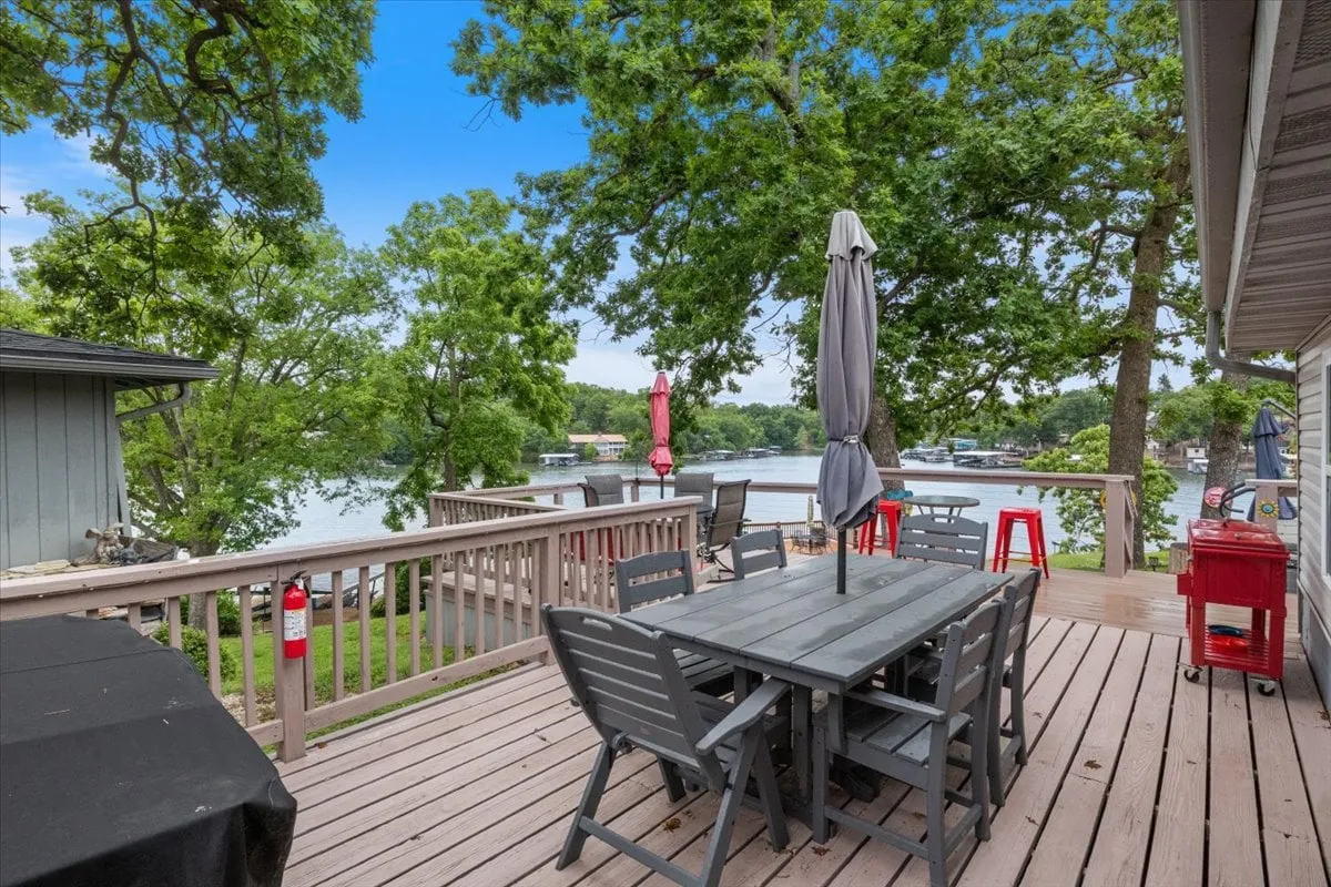 This exterior image shows a spacious deck with a large dining table, an umbrella, red accents, and a stunning view of the lake framed by trees. It’s set up for outdoor dining and socializing, seamlessly connecting indoor and outdoor living.