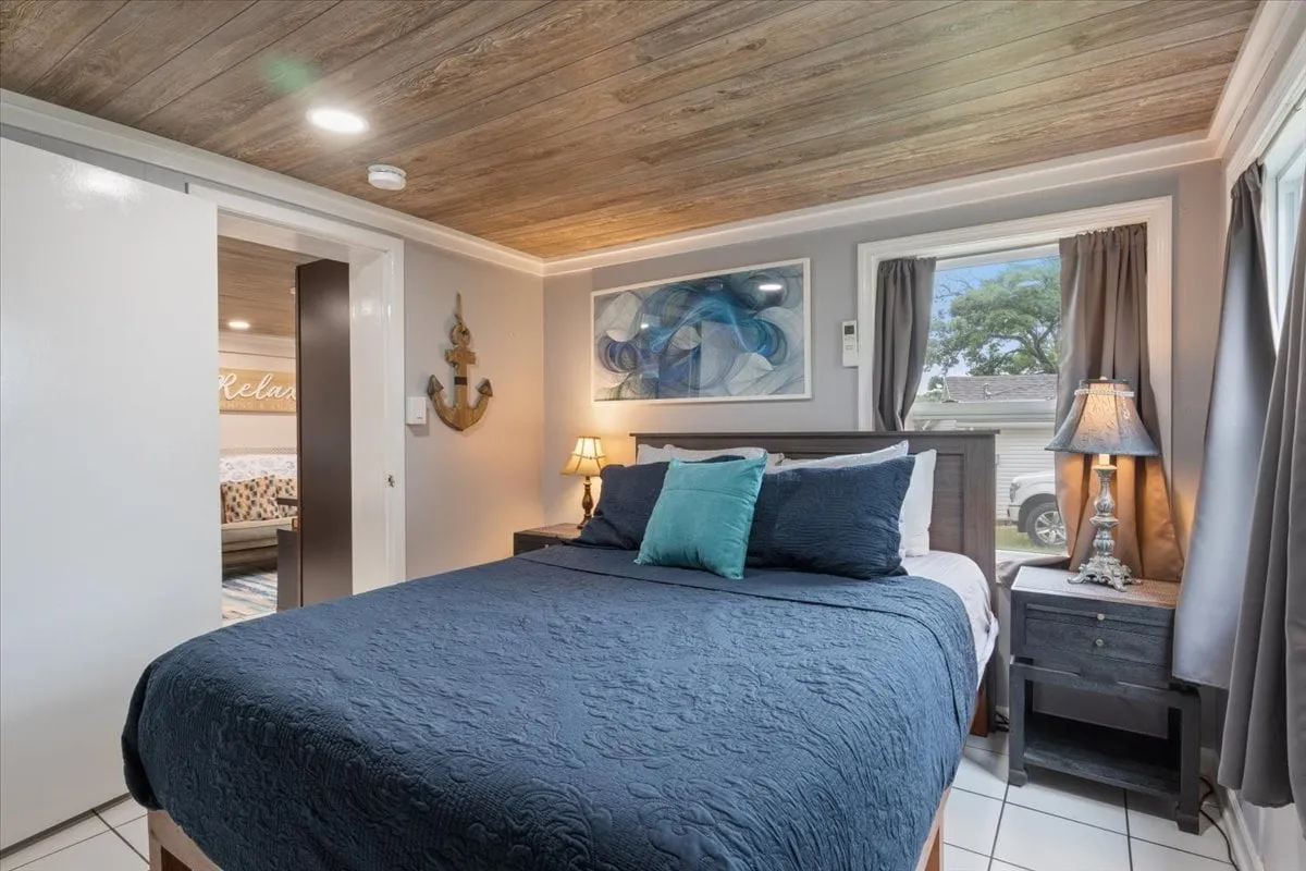 A serene bedroom with a comfortable bed dressed in dark bedding and blue accent pillows. The room features elegant wall art, soft table lamps, and a window that fills the space with natural light. The overall ambiance is calm, cozy, and inviting—perfect for unwinding after a day at the lake.