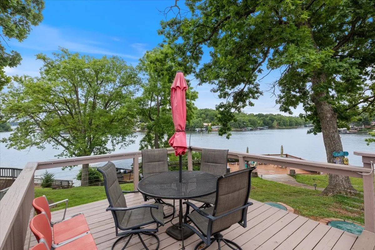 A serene outdoor dining area under a pink umbrella, with a direct view of the lake and surrounded by green trees. The setting is calm and welcoming, ideal for quiet moments or socializing with companions—including pets.