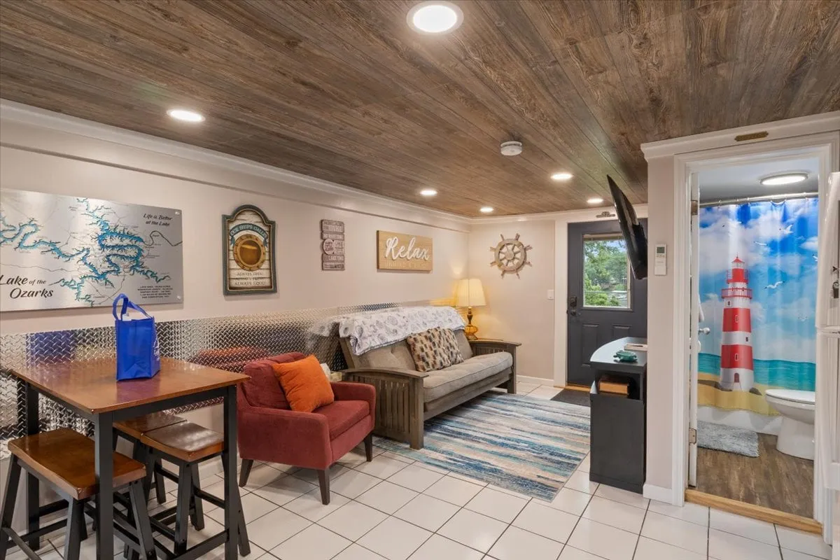 A welcoming living room featuring a sofa with a decorative throw, a small table and chairs, and a hint of a lighthouse-themed shower curtain in the background. The area is cozy and multifunctional, hinting at flexible sleeping arrangements like a pullout couch.