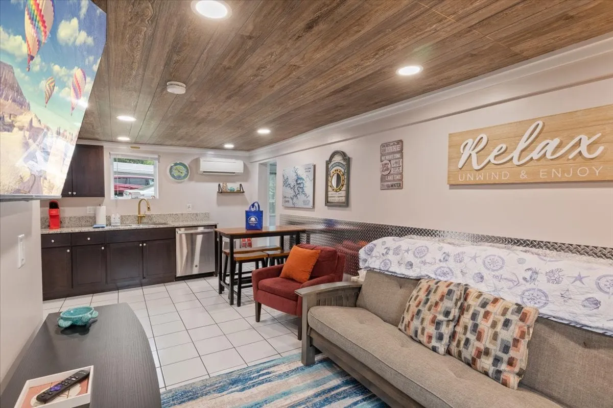 This image captures an inviting open-plan living area with a sofa adorned with decorative pillows, a modern kitchen area with updated appliances, a dining table, and cheerful wall art including a 'Relax' sign. The space is bright and airy, suggesting an easy flow between lounging, dining, and cooking areas.