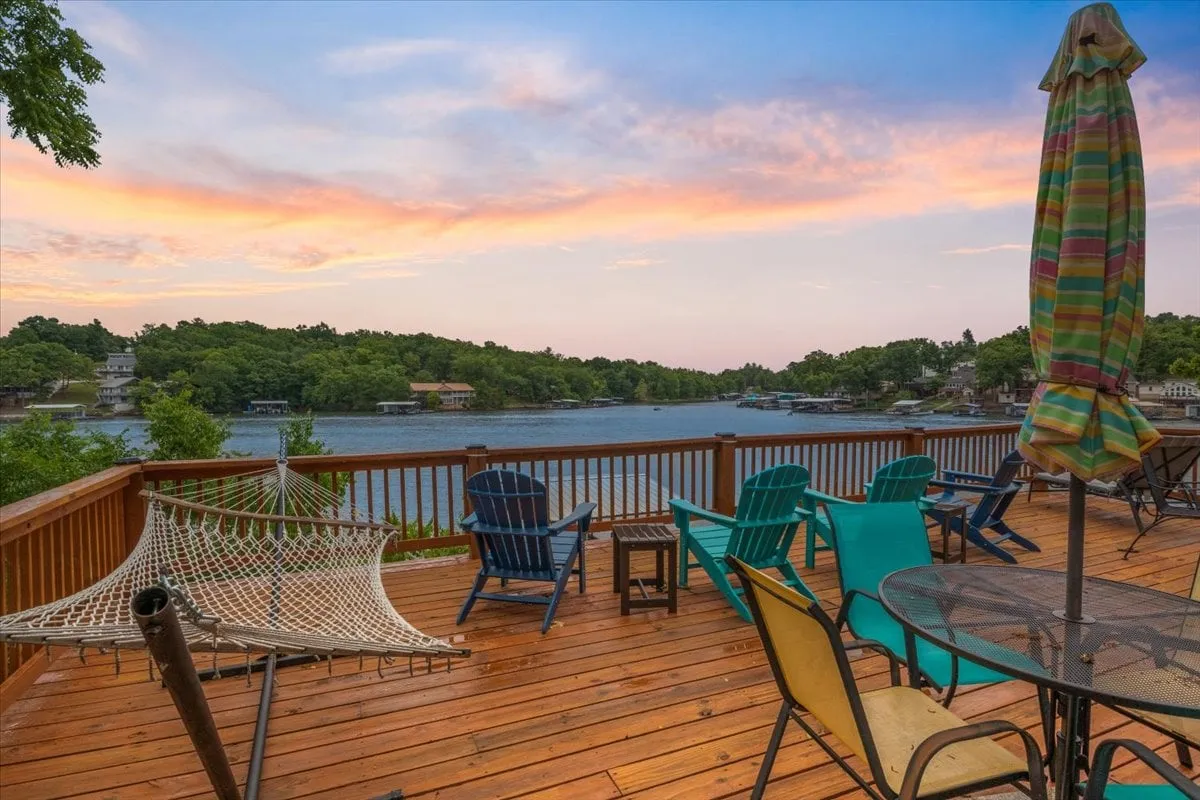 The image showcases a sun-drenched lakeside deck at Hawk's Landing, complete with a hammock gently swaying in the breeze, comfortable deck chairs, a shaded table with an umbrella, and sweeping views of the tranquil lake—perfect for relaxation, outdoor dining, and taking in the sunset.