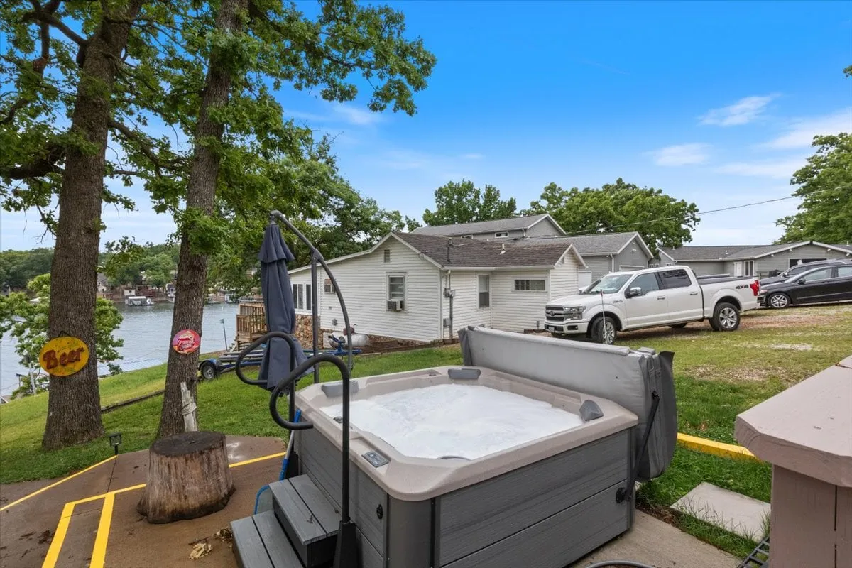 This inviting exterior shot showcases a lakeside hot tub with surrounding trees, lake views, and outdoor seating. The setup reflects a space designed for both relaxation and group gatherings, with the lake easily accessible for swimming and boating.