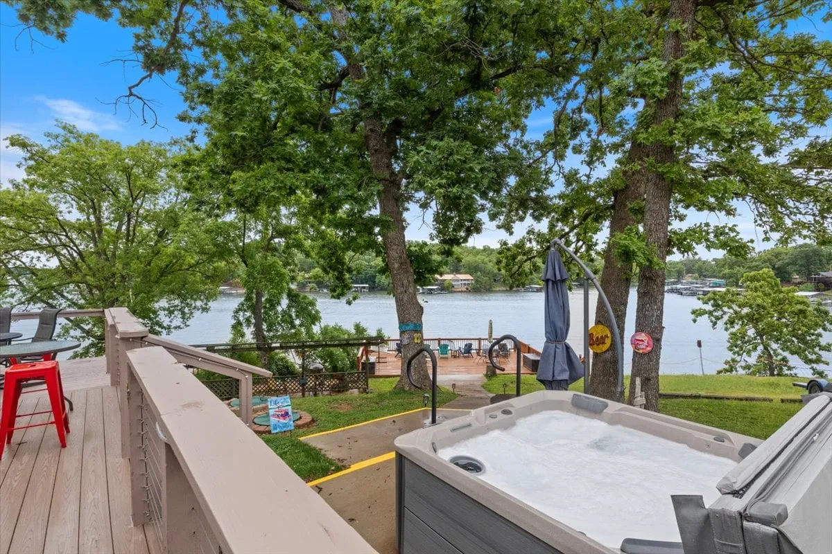 Expansive deck overlooking the lake, featuring a hot tub with panoramic views, comfortable seating, and shaded areas beneath tall trees. The lake stretches out beyond the deck, inviting guests to relax and soak up the scenery.