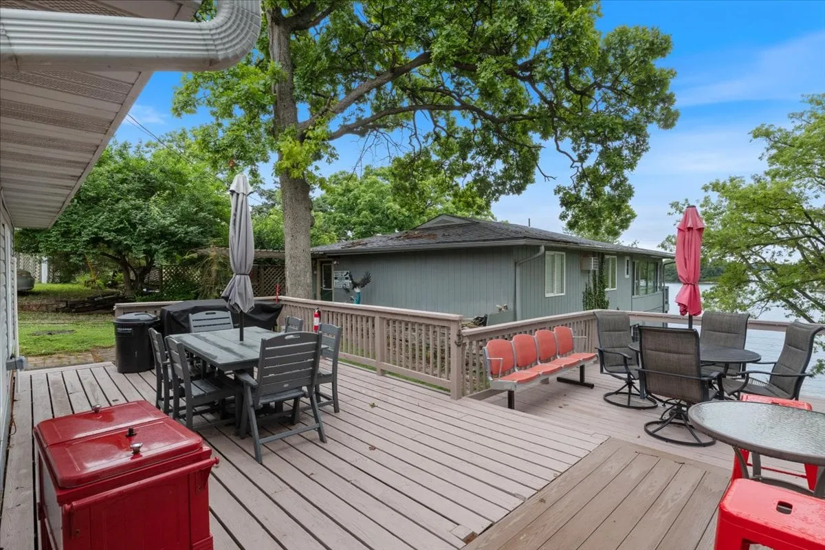 A spacious wooden deck with an outdoor dining set, red cooler, and patio umbrellas, overlooking the water. The area is lively, inviting, and perfectly suited for gatherings or unwinding outdoors with pets nearby.